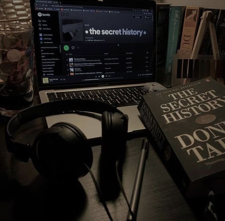 the image shows a dimly lit desk with a laptop open to a spotify playlist titled "the secret history." a physical copy of the book "the secret history" by donna tartt sits next to the laptop. headphones and a pen are also on the desk, along with a glass and bookends holding books. the scene evokes a dark academia aesthetic.