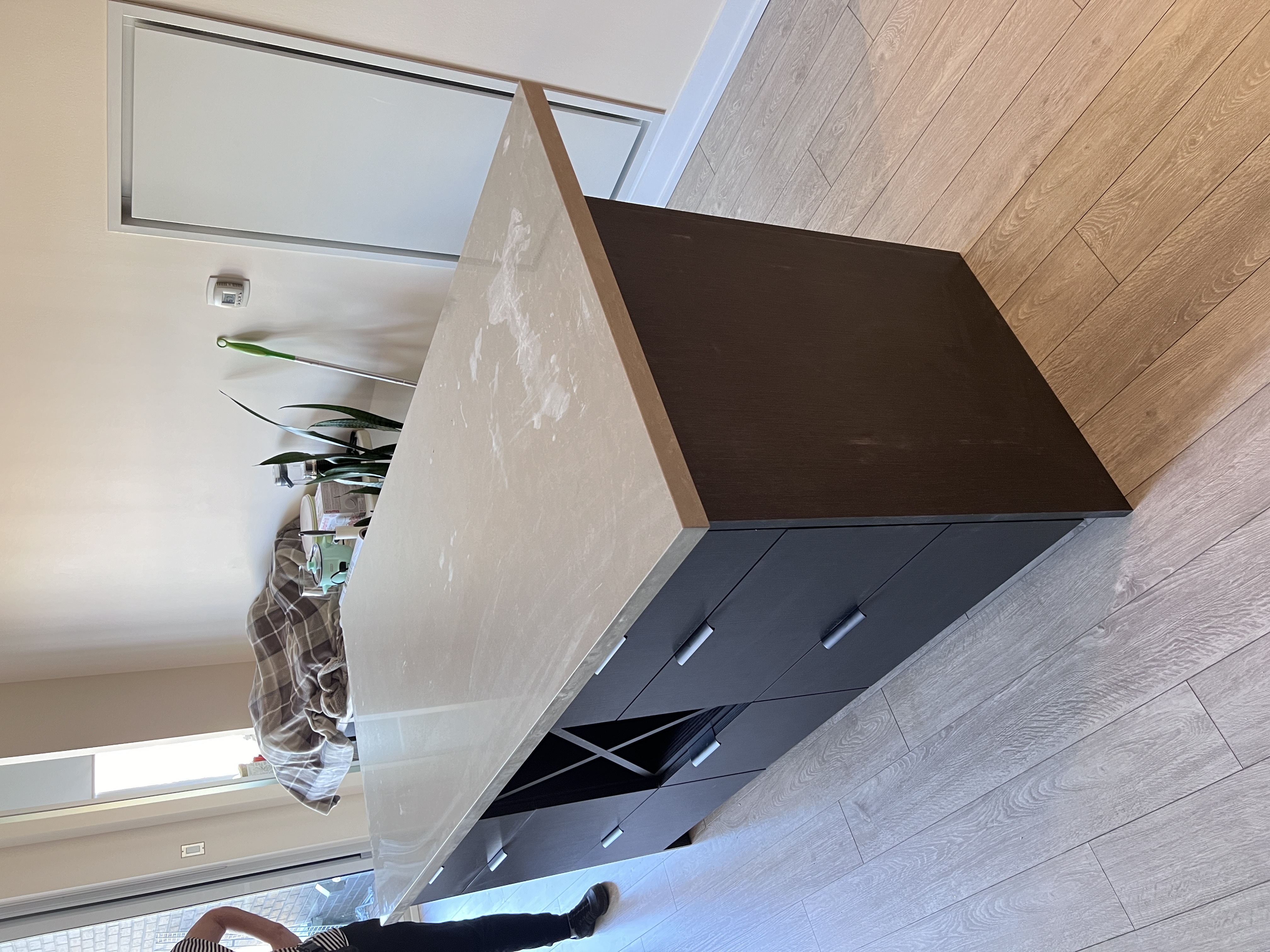 he image shows a modern kitchen island with a light gray concrete countertop and darkcolored cabinets. the island features several drawers and a builtin wine rack. the flooring is light gray laminate. a portion of a person's leg is visible in the background, and there is some household clutter visible behind the island.  the overall style is modern and minimalist.