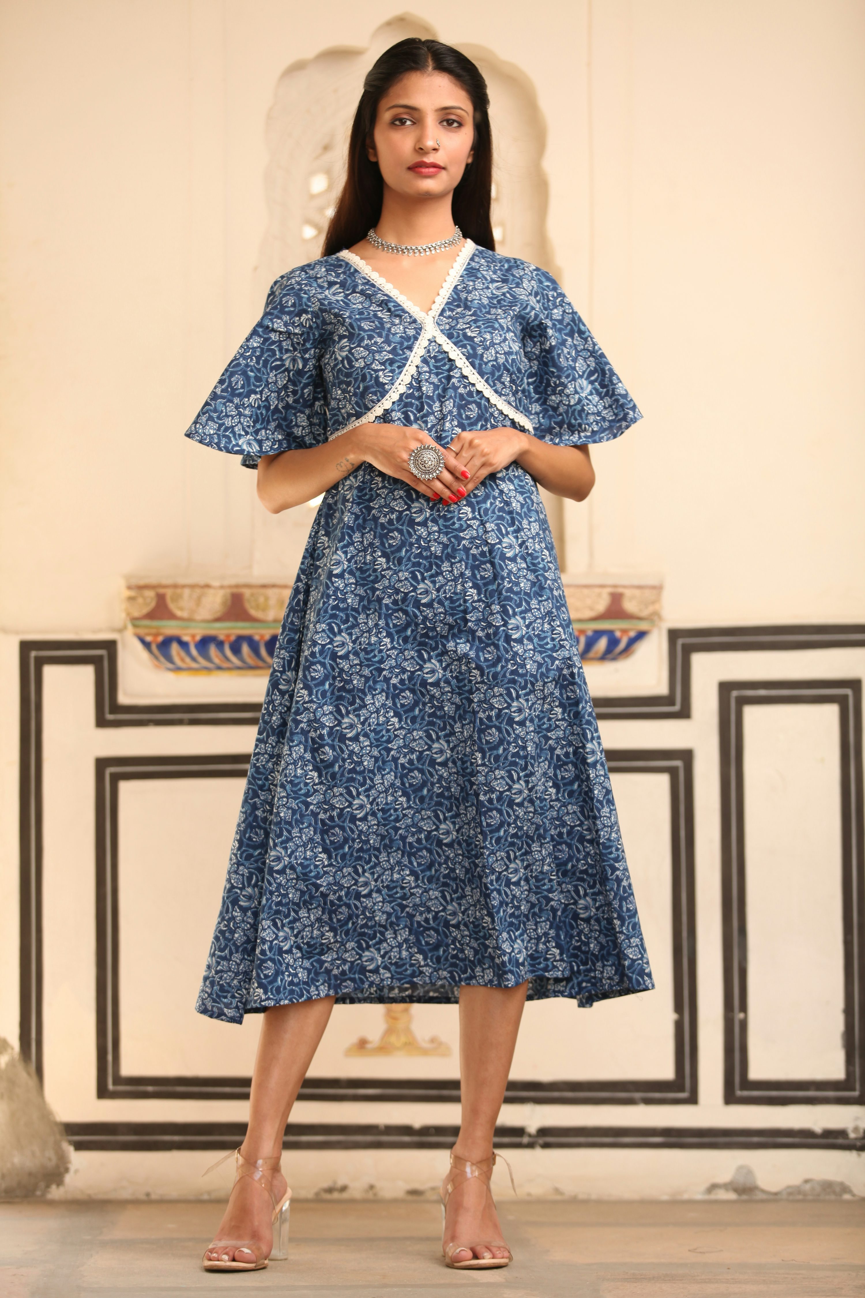 a fulllength shot of a young indian woman wearing a beautiful blue floral midi dress. the dress has bell sleeves, a vneck with a crossover detail, and is adorned with white lace trim. the dress is made from a light cotton material and has a delicate indigo block print. she is accessorized with a silver necklace and ring, and wears clear high heels. the background is a traditional indian setting with ornate wall decor. the image is professionally shot, showcasing the dress and the model’s style.