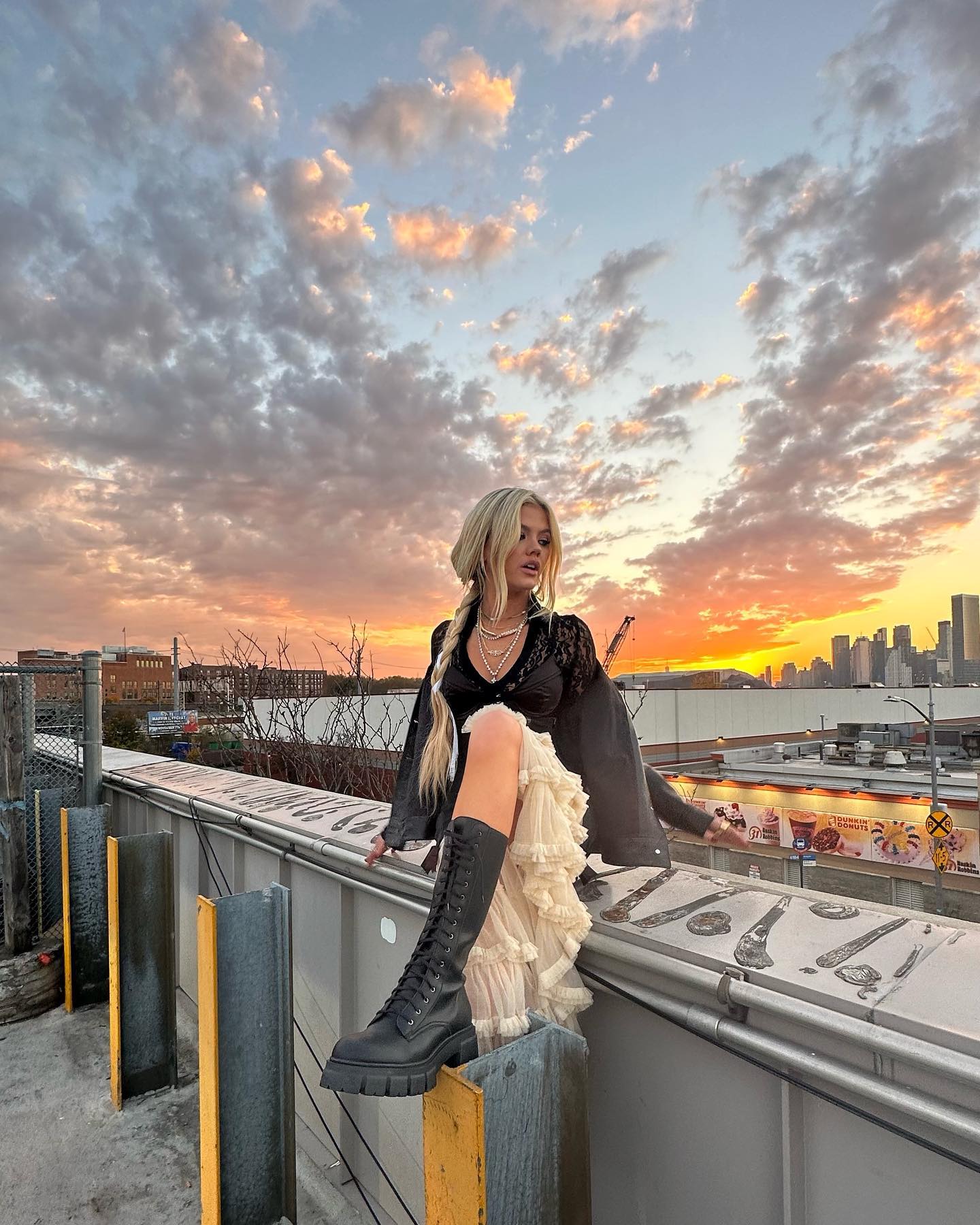 a blonde woman sits on the edge of a rooftop at sunset. she is wearing a black lace top, a cream ruffled skirt, and tall black boots. her hair is braided and she is wearing a necklace. behind her is a vibrant sunset with pink and orange clouds over a distant cityscape. a fence and some industrial structures are visible in the foreground.