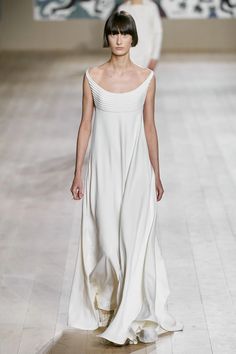 a model wearing a long, white dress walks down a runway at a fashion show. she is posing for the cameras and looking elegant and stylish. her hair is short and she has a beautiful smile. the dress is a simple, but elegant design, with a high neckline and a flowing skirt. it is a perfect example of how simple and beautiful fashion can be.