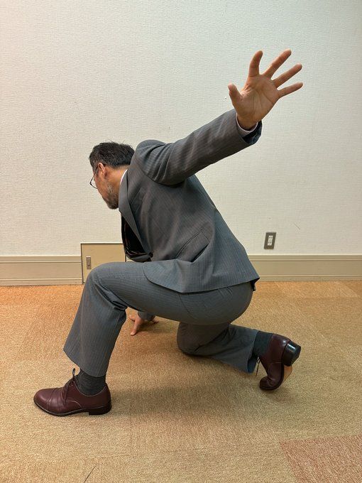 a man in a gray business suit is dramatically crouching and reaching towards the left side of the image. he is in an office setting with plain walls and carpeted floor. the lighting suggests an indoor environment.