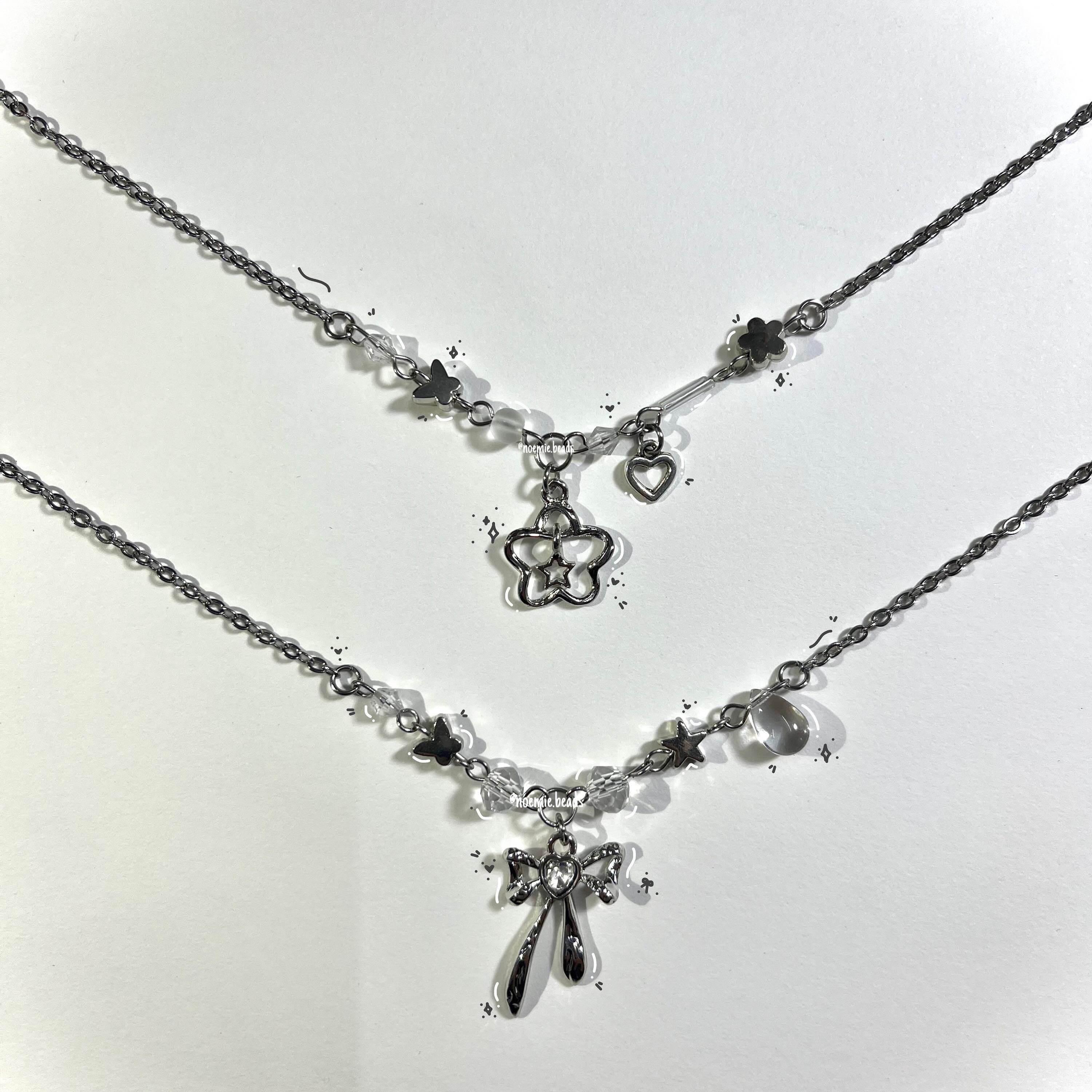 two silver necklaces lie on a white surface. the top necklace features a silver chain with butterfly, flower, and heart charms and a flower pendant. the bottom necklace features a silver chain with butterfly, star, and clear bead charms and a bow pendant.