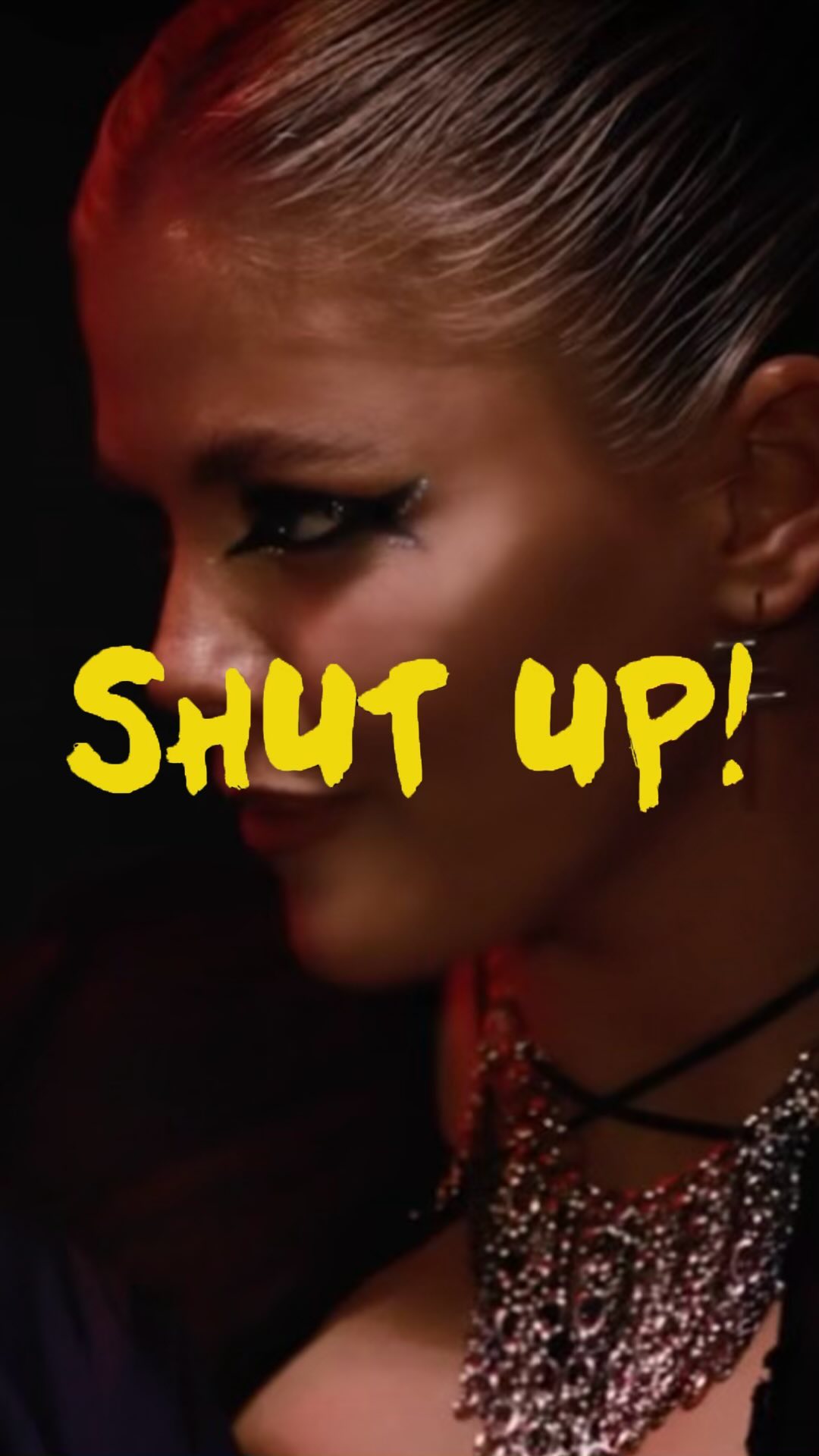 a closeup shot of a woman with dramatic makeup and edgy jewelry. the text "shut up!" is superimposed in yellow over her face.