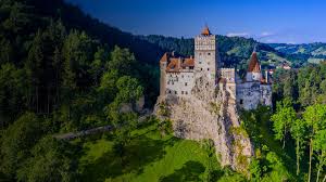 the image showcases a majestic medieval castle, bran castle, perched atop a hill and enveloped by lush greenery. bathed in the warm glow of a sunny day, the castle, with its pointed towers and stone walls, stands as a testament to history. the surrounding forest, vibrant and verdant, stretches out as far as the eye can see. the clear blue sky dotted with fluffy white clouds provides a serene backdrop to this captivating scene. the aerial perspective offers a breathtaking view of the castle and its picturesque surroundings.
