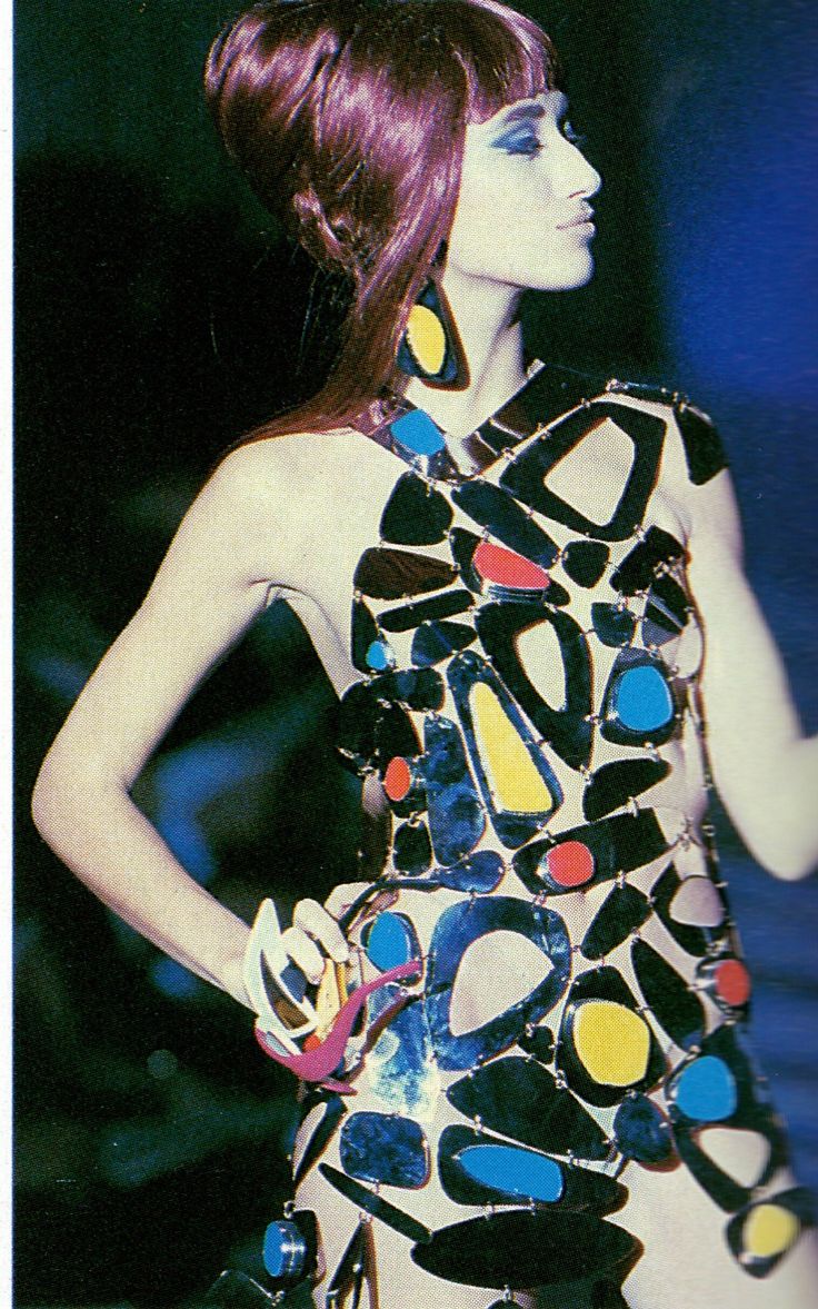 a fashion model is shown in a photograph wearing a dress made of irregularly shaped metallic pieces with colorful accents. she also has large earrings, a bracelet and an elaborate updo hairstyle with bangs. her makeup includes heavy eye makeup. she is posing in a profile view.