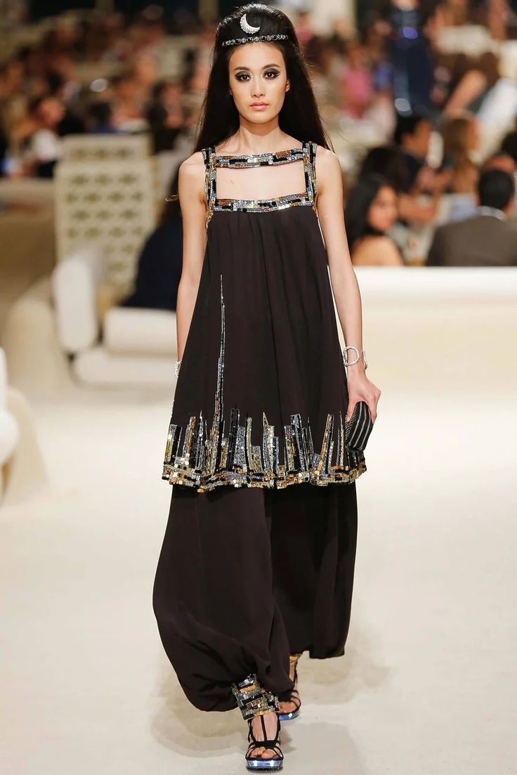 a fashion model walks the runway in a black dress with a city skyline design made of sequins. she is wearing highheeled sandals and her hair is styled in an updo with a hair clip. she looks confident and stylish. the dress is long and flowy, and the sequins shimmer in the light.  the model is wearing jewelry and accessories, including a watch on her wrist and a ring on her finger. the background is a white backdrop with a light pattern. this image is a great example of high fashion and couture.