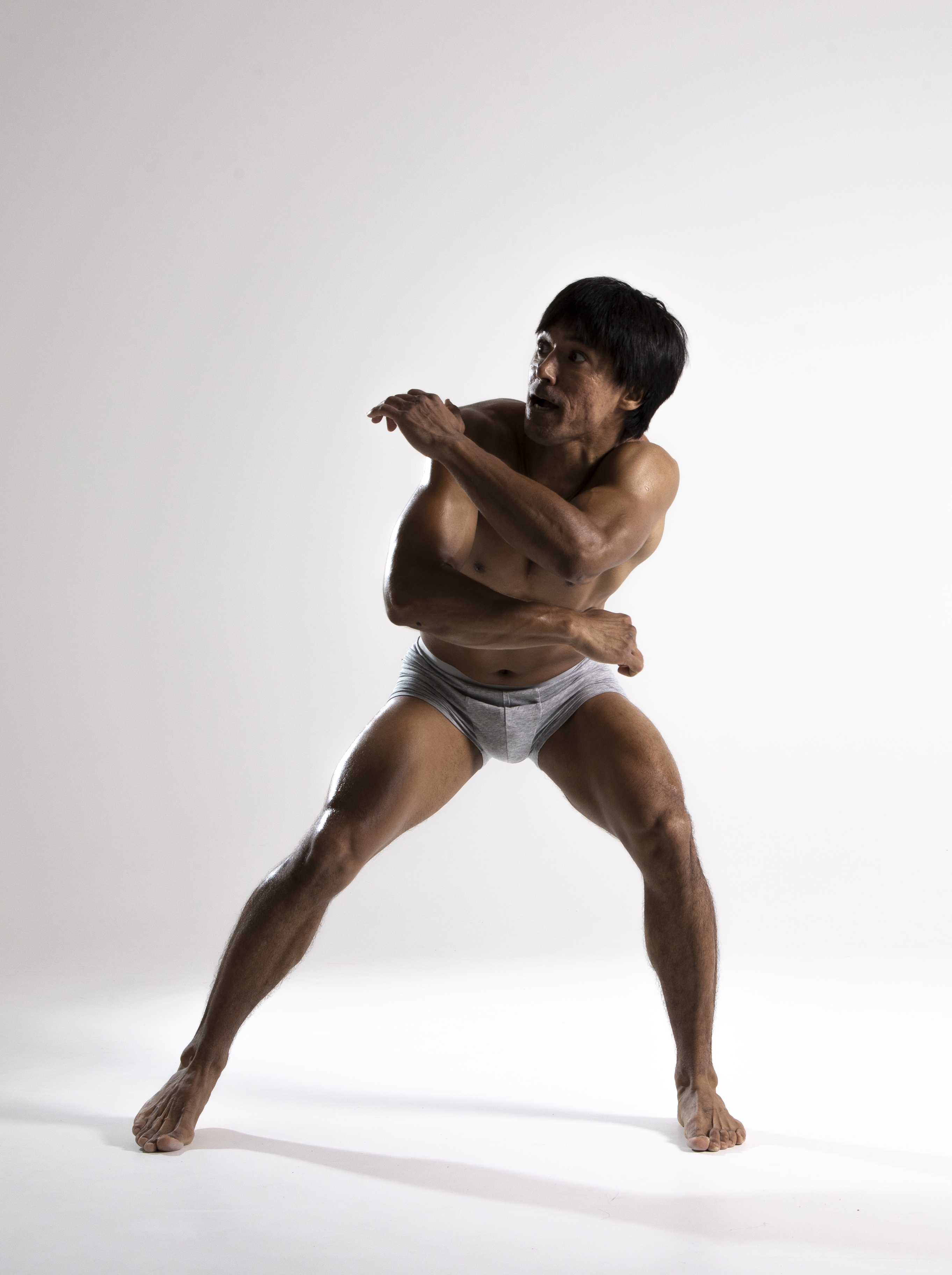 a muscular, barechested man in gray underwear poses confidently against a white background. he stands with legs spread and arms crossed, looking away with a determined expression. his physique is toned and athletic, suggesting fitness and strength. the studio lighting highlights his muscles and contours.