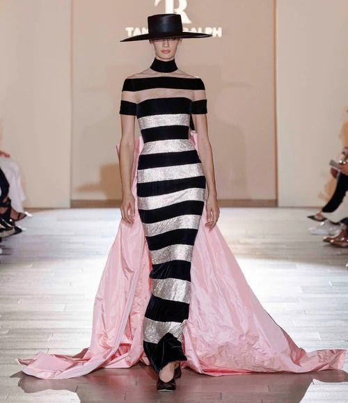 a model walks the runway wearing a black and white striped dress with a pink train. she is wearing a black hat and black shoes. she is posing for the camera with her left hand on her hip.