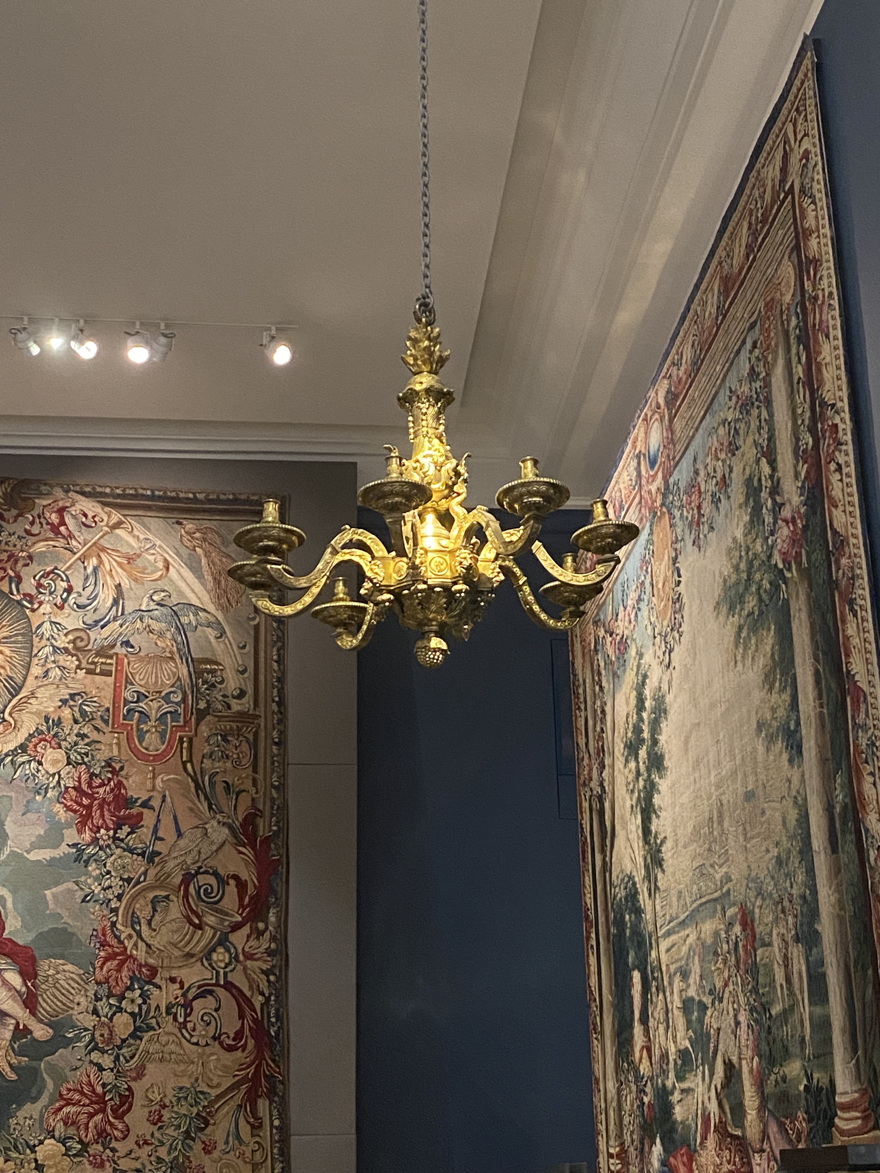 the image shows a gold chandelier hanging in a museum. behind the chandelier are two large tapestries hanging on the walls. the walls are painted in different colors, and the ceiling has recessed lighting. the overall scene is ornate and suggests a historic or artistic setting.