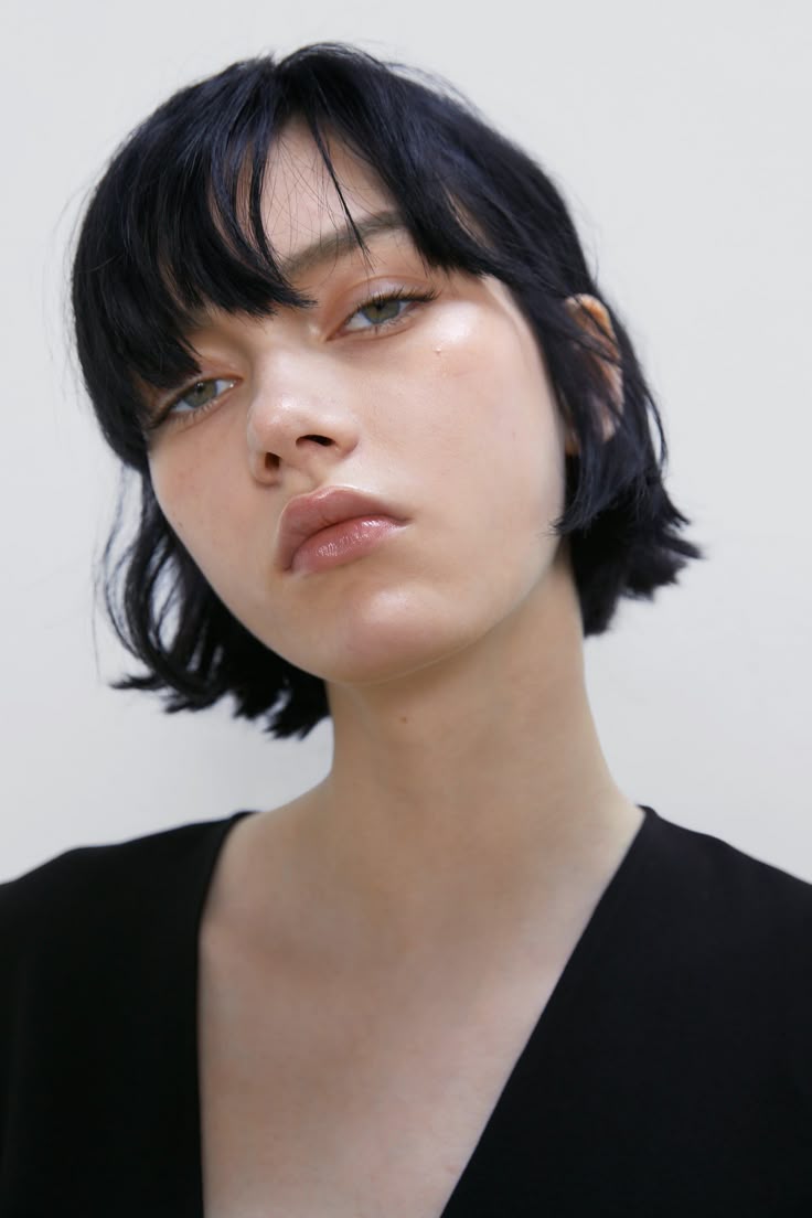 a closeup portrait of a young woman with short, dark hair and bangs. she has fair skin, green eyes, and glossy lips. she is wearing a black top with a deep vneck. her hair has a slightly wet or styled texture.