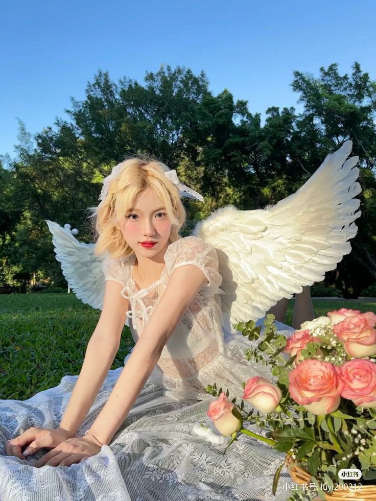 a young woman with blonde hair and angel wings sits on a lace picnic blanket outdoors. she is wearing a white lace dress and a headband. next to her is a basket filled with pink roses. the background features a green lawn, trees, and a clear blue sky. she has fair skin and is wearing red lipstick.