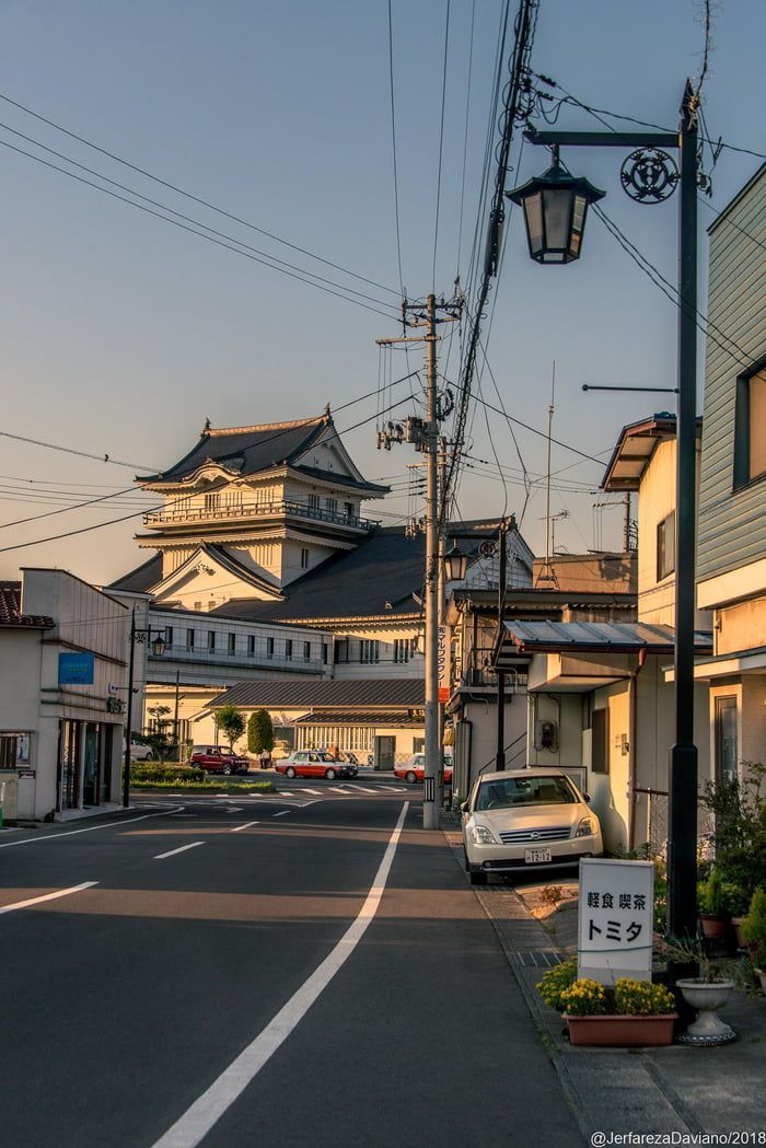 a tranquil street scene in japan, possibly a smaller town or city, is presented. the focal point is a traditional japanese building, reminiscent of a castle or fortified structure, in the background. the foreground features a quiet street with parked cars, lined by traditionalstyle houses.  overhead power lines are prominent, and a streetlamp casts a soft light. the overall atmosphere is serene and peaceful, characteristic of a quiet evening in a japanese town. the image has a calm, almost nostalgic quality, capturing the everyday charm of the location. a small sign in japanese is visible near the foreground.