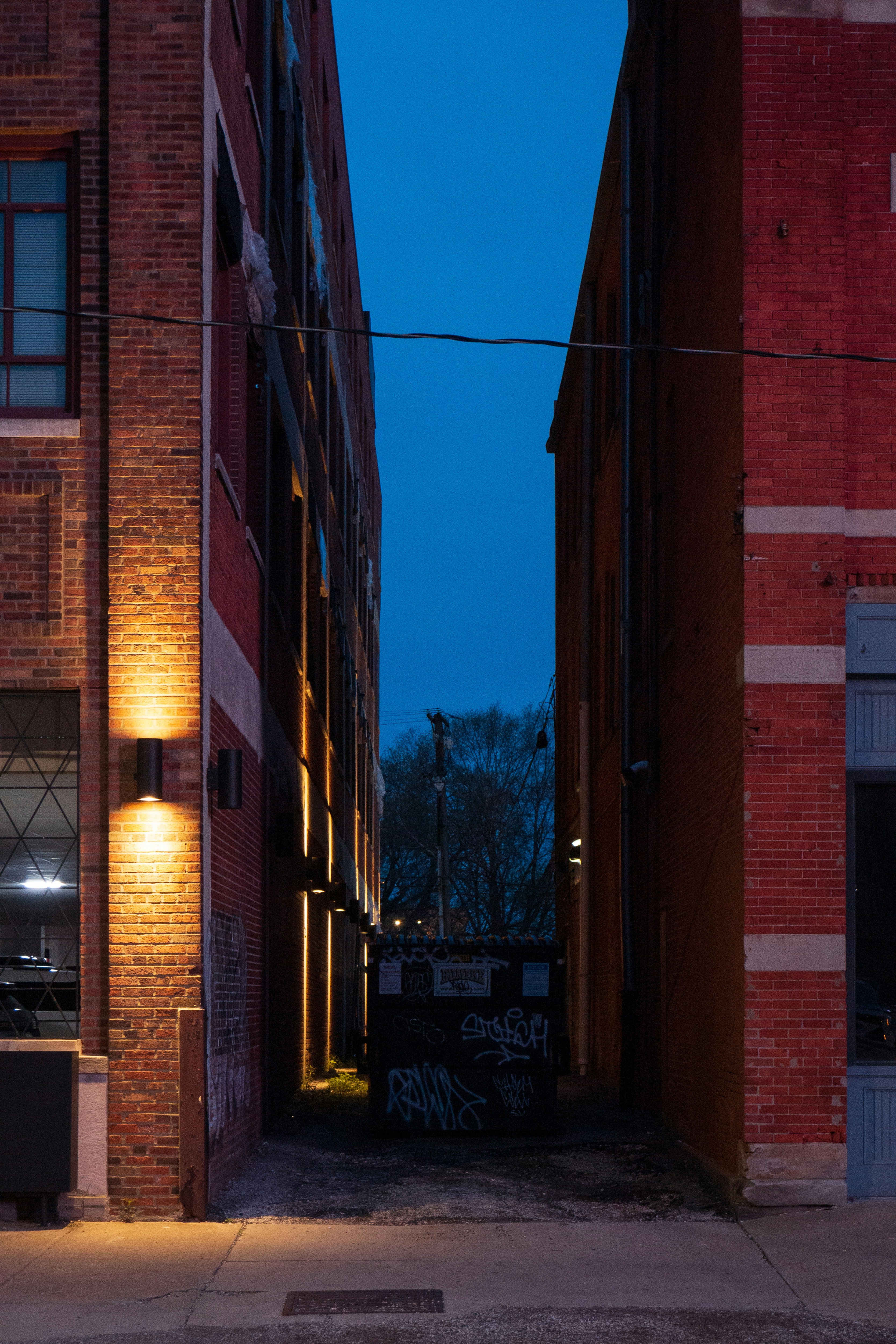 the image shows a narrow alleyway between two brick buildings at night. the buildings are lit by streetlights, and there is a dumpster in the alley. the sky is dark blue.