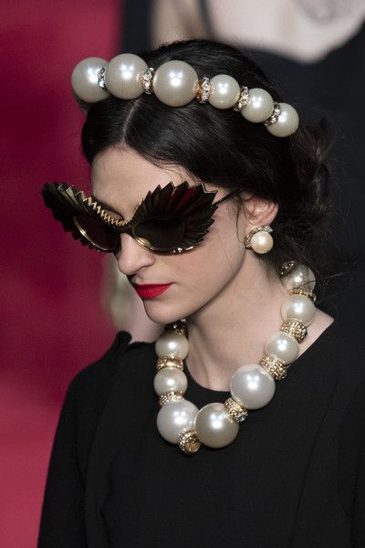 a model wearing a black dress walks the runway. she has a pearl headband, large pearl necklace, large sunglasses with gold accents and large pearl earrings.