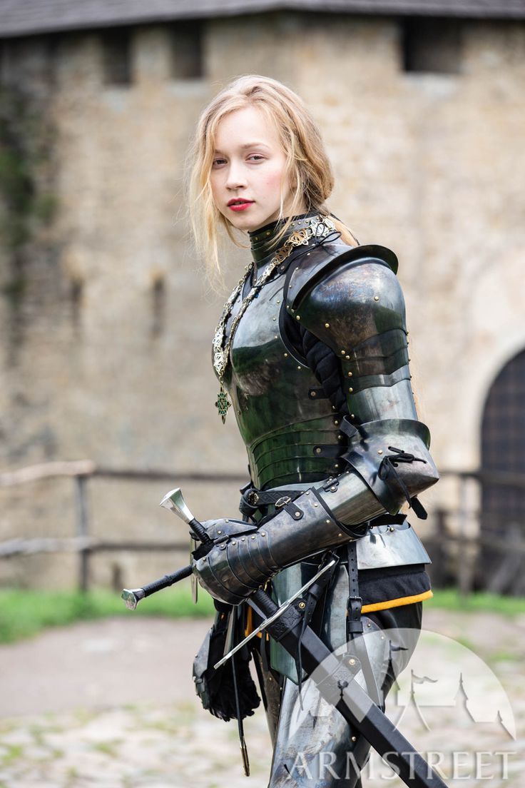 a woman in full medieval armor poses with swords in front of a stone building. she has blonde hair and wears red lipstick. the armor is shiny and black, with intricate details around the neck. she holds a sword in each hand, and additional swords are attached to her belt. the building in the background appears to be a castle or fortress. the overall impression is one of strength, history, and a powerful female warrior.