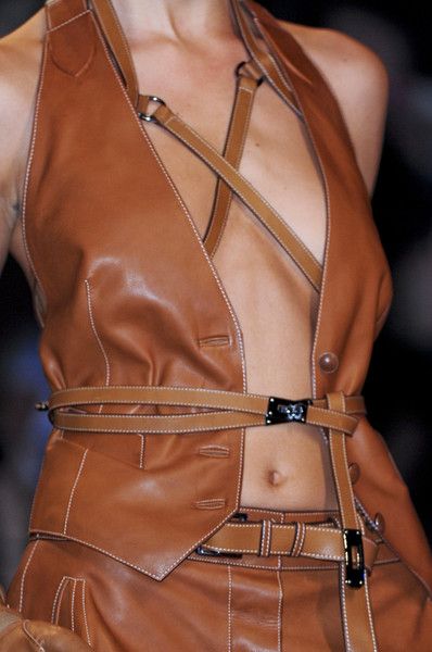 a woman model wearing a  leather jacket and vest combo with a  brown leather belt around her waist,  showing a toned midriff.  she is posing on a runway in a fashion show.  the outfit is styled in a  contemporary,  and edgy way.