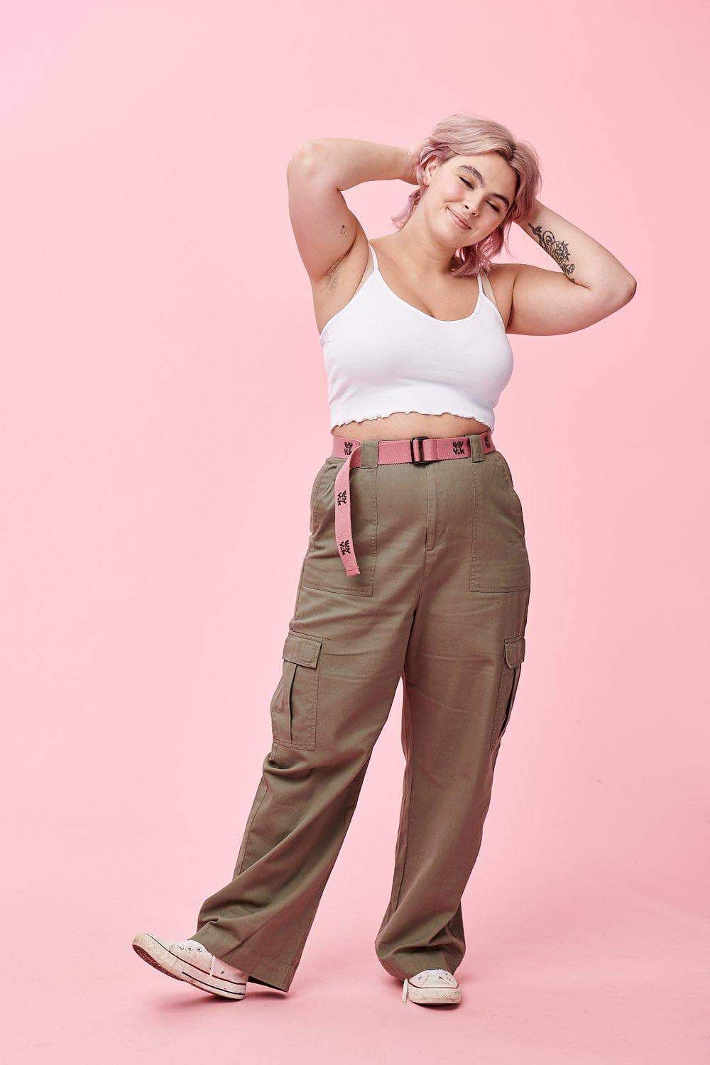 a young woman with pink hair  stands confidently against a pastel pink backdrop.  she's styled in a white crop tank top and highwaisted olive green cargo pants. a pink belt with a black buckle cinches her waist. her pose is relaxed and natural, with one leg slightly extended and her hands casually placed behind her head. she's wearing white sneakers, completing the casual and trendy look.