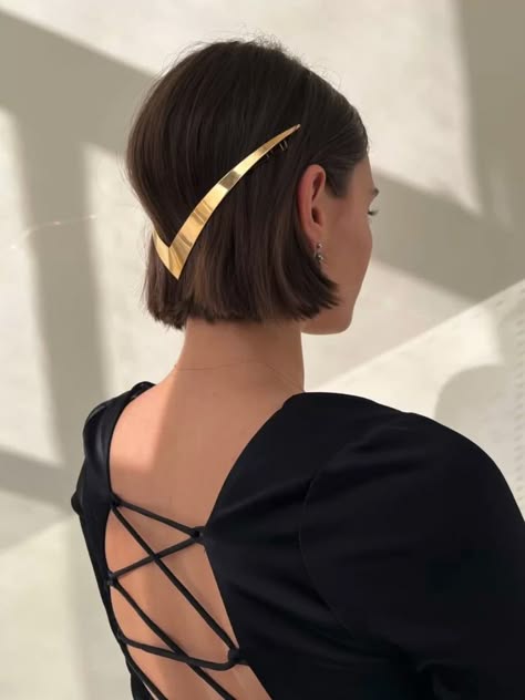 a woman with short bob hair, styled with a gold hairpin, is wearing a black dress with a laceup back. her hair is styled in a chic and modern way, with the hairpin adding a touch of elegance. she is looking away from the camera, creating a mysterious and alluring image. the overall look is stylish, sophisticated, and elegant.