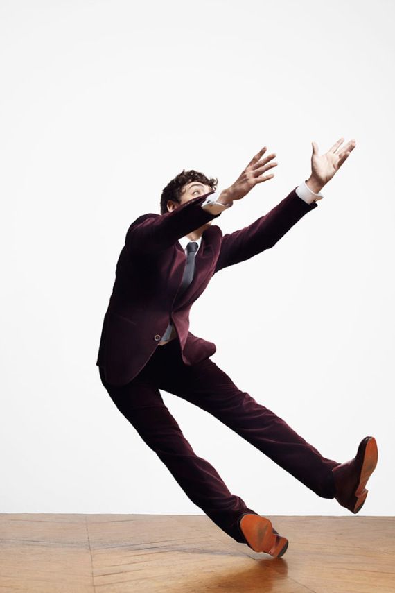 a man in a maroon velvet suit is captured midair, executing a dynamic jump or dance move against a clean white studio backdrop. he is leaning back with his arms outstretched, creating a sense of energy and motion. the low camera angle emphasizes the height and dynamism of his pose. the lighting is bright and even, highlighting the texture of his suit and the sleekness of his movements.