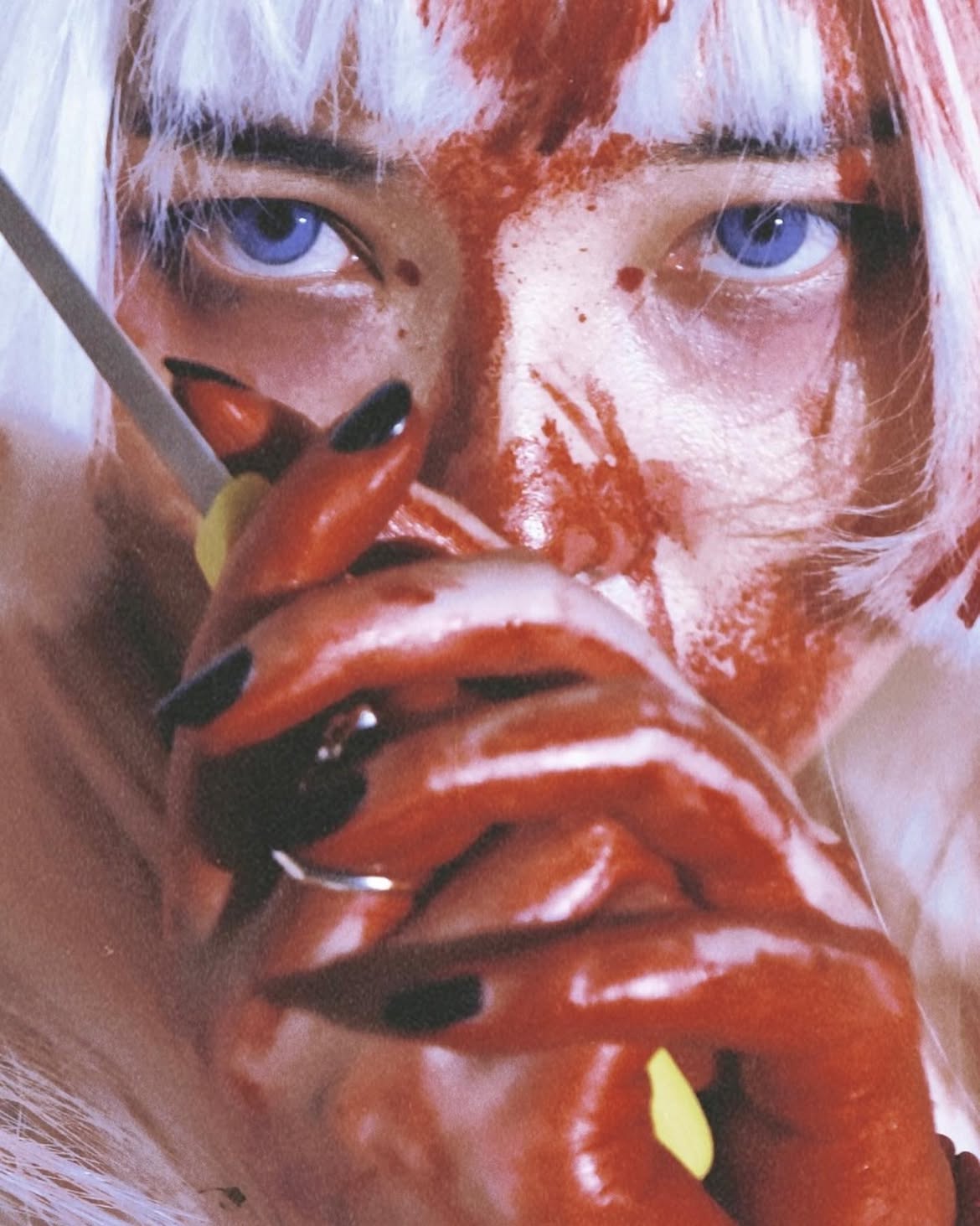 the image shows a person's face covered in what appears to be blood. they have white hair and striking blue eyes. their hands, also covered in blood, are near their face, and one hand holds a cigarette. they have dark nails. there is a ring on one of their fingers.