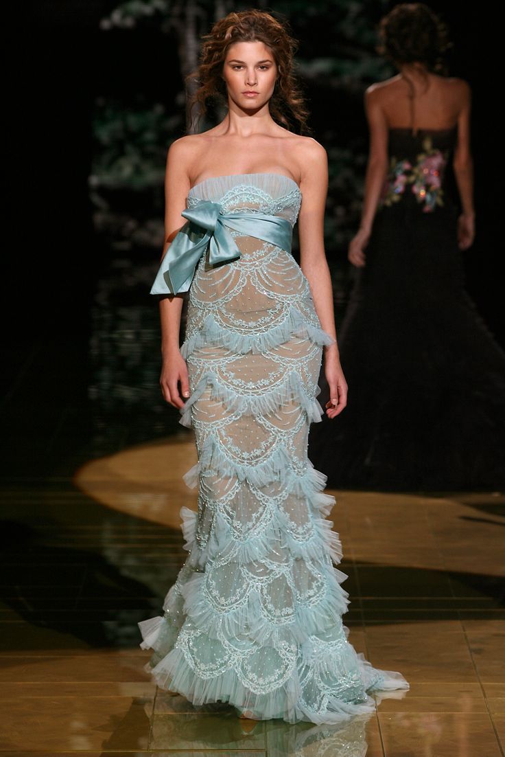 a model walks the runway during a fashion show wearing a light blue strapless gown with layers of embroidered tulle and a large blue ribbon at the waist. another model is partially visible in the background wearing a black strapless gown with floral embroidery.