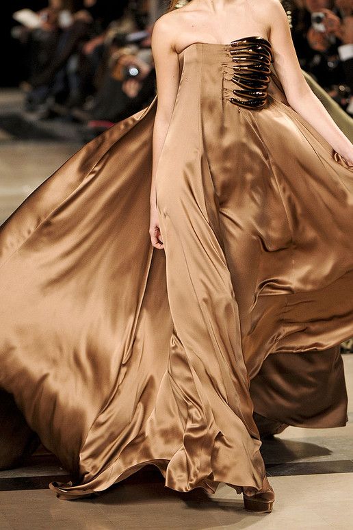 a woman wearing a long strapless brown satin dress on a runway. the dress has a unique design on the bodice and flows dramatically as she walks. the background shows a blurred crowd of people.