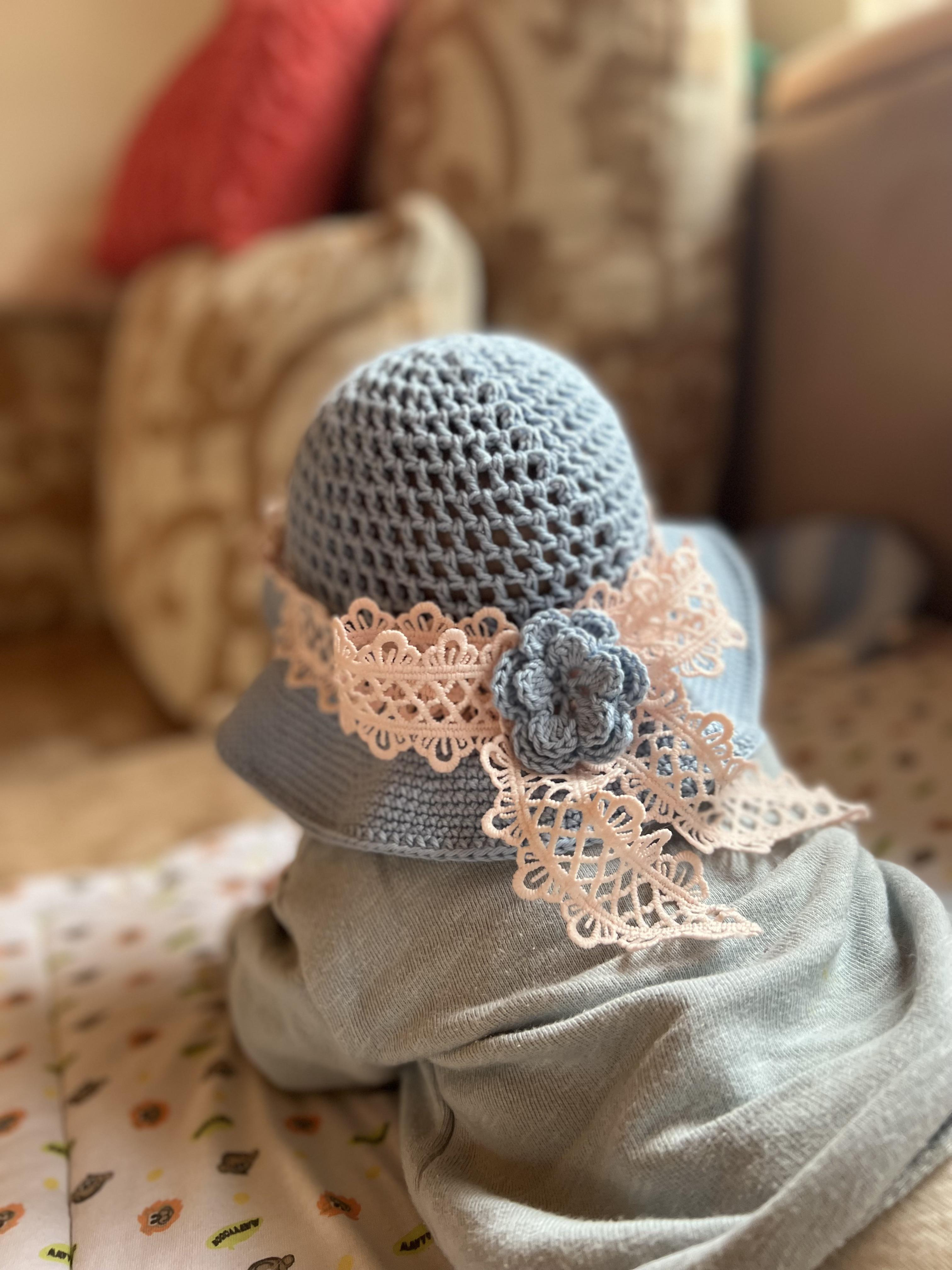 loseup view of a baby wearing a light blue crocheted sun hat adorned with delicate pink lace. the hat is intricately crafted, and the lace adds a touch of femininity.  the baby is sitting on a lightcolored changing mat or blanket with a patterned design, partially visible at the bottom of the image.  the background is blurred but shows a couch with several pillows, suggesting a home environment. the overall feel of the image is warm, soft, and evokes a sense of calm.