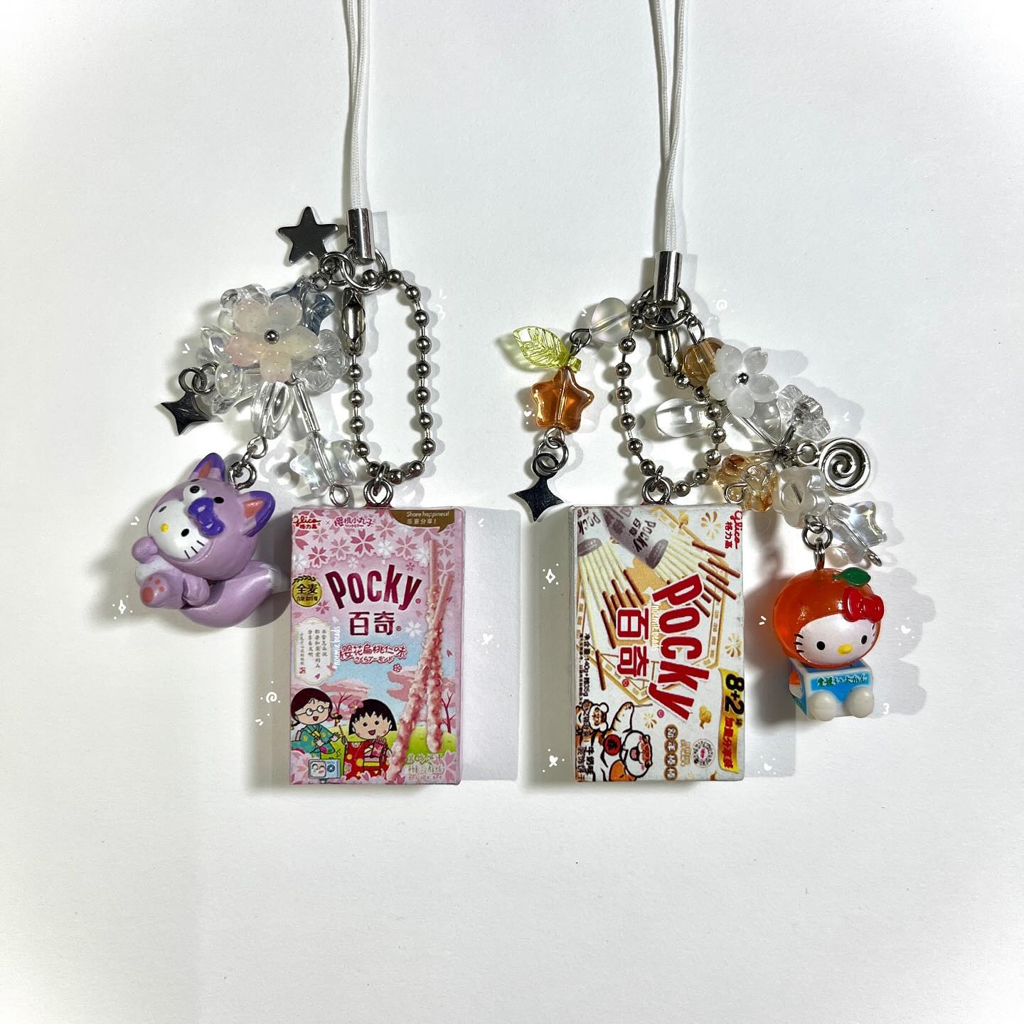 two cell phone charms are displayed. each charm features a mini pocky box, hello kitty figure, beaded chain, and a variety of star, flower, and transparent beads. the charm on the left has a purple hello kitty figure and a pink pocky box. the charm on the right has an orange hello kitty figure and a brown pocky box.