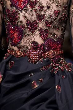 a closeup image of a woman's dress that is a rich dark navy and completely covered in red jeweled hearts. the dress is embellished with a shiny black lace and the bottom half appears to be made of a flowing satin fabric.  the model is standing in a darkened room, with her bare shoulders showing. this luxurious dress is glamorous and perfect for a special occasion.