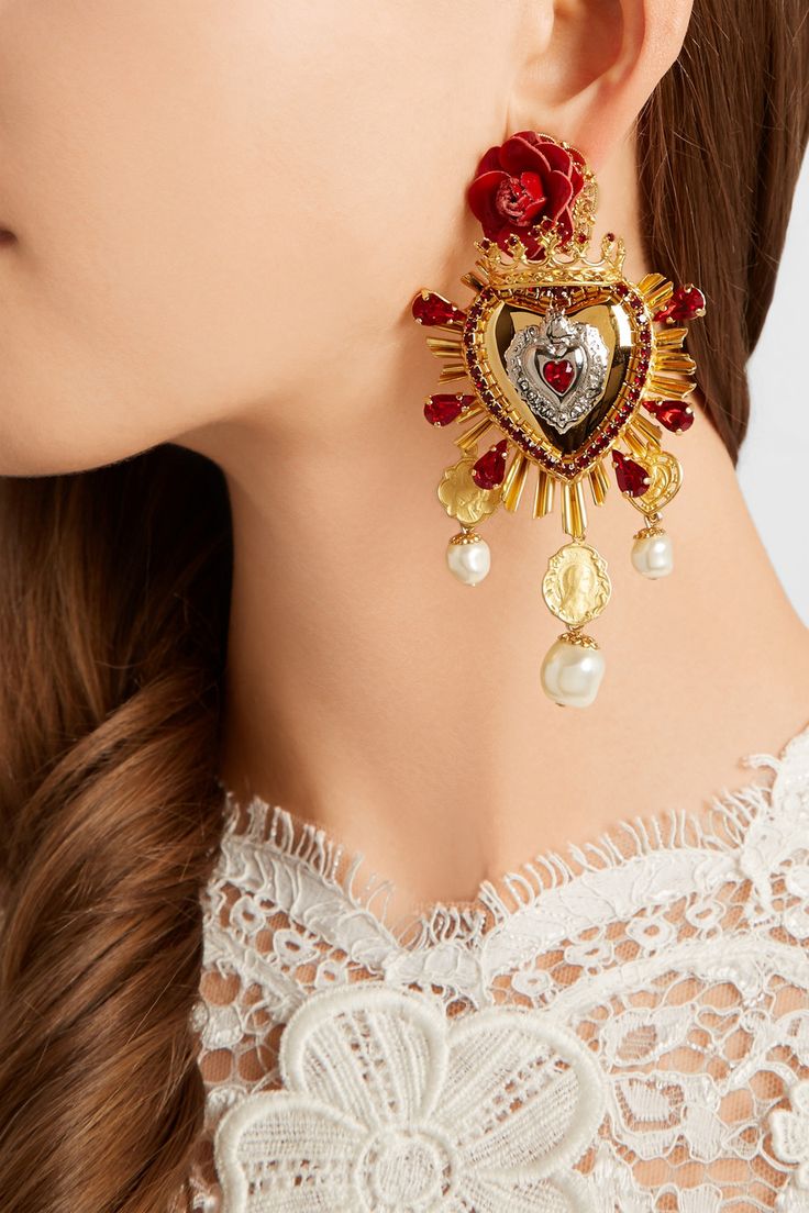 closeup shot of a woman wearing a pair of large, ornate dolce & gabbana earrings. the earrings are gold and feature a red heart design with pearls and roses, with religiousinspired details.  the woman is wearing a white lace top. the image is focused on the earrings and provides a detailed view of their intricate design.