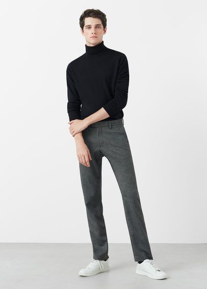a fulllength studio shot features a young man in a black turtleneck sweater, gray trousers, and white sneakers. he stands against a plain white background. his left arm is bent across his torso, and his right arm hangs at his side.