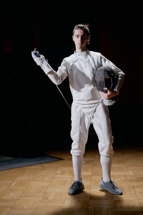 a young male fencer stands on a polished wooden floor, holding his foil and mask. he's wearing a white fencing uniform and grey shoes. the background is dark, focusing attention on the athlete. the image portrays a sense of readiness and focus.