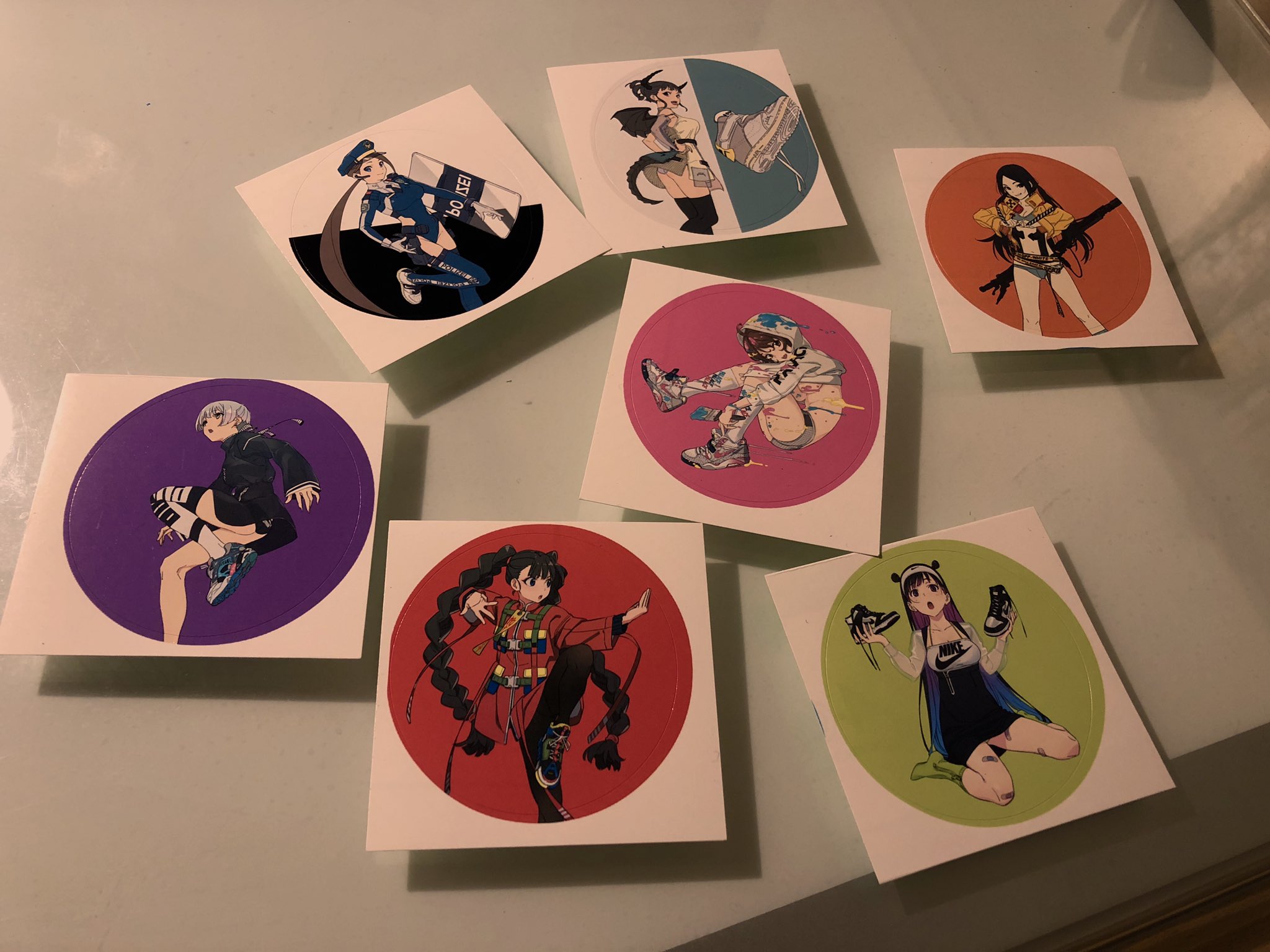 the image shows seven square stickers arranged on a table. each sticker features a dynamic illustration of a female anime character, often in midaction poses. the characters have distinct outfits and hairstyles, suggesting they may be from the same anime or game. the stickers are set against bright, solid color backgrounds that contrast with the character art.