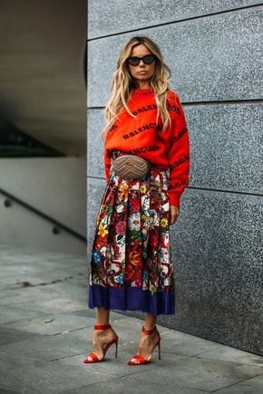 a stylish blonde woman is standing on a city street. she is wearing a red balenciaga logo sweater, a colorful floral midi skirt, a gucci belt bag, and red high heels. her look is chic and trendy, showcasing a mix of highend and highstreet brands. the background is a modern urban setting with a gray stone wall. the overall image exudes confidence and a sense of fashionable street style.