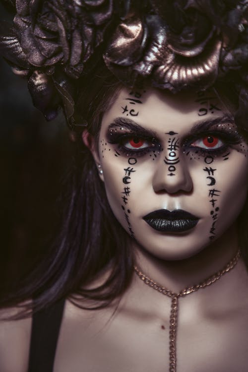 loseup portrait of a woman with elaborate dark makeup. she is wearing a dark flower crown, and her face is adorned with dark symbols that resemble runes or occult markings. her eyes are a striking red, and her lips are painted black.  she is wearing a gold chain necklace. the overall mood is dark, mysterious, and slightly spooky, suggesting a gothic or fantasy theme.