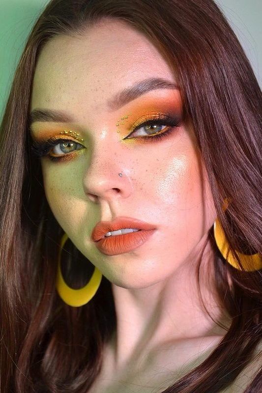 a closeup portrait of a woman with elaborate orange eyeshadow, yellow hoop earrings, and a nose piercing. she has a fair complexion with freckles, and her hair is brown and styled in loose waves. her makeup is meticulously applied, featuring bright orange shades on her eyelids and under her lower lash line, accentuated with small, shiny embellishments near the inner corners of her eyes. her lips are colored with a matte orange lipstick.
