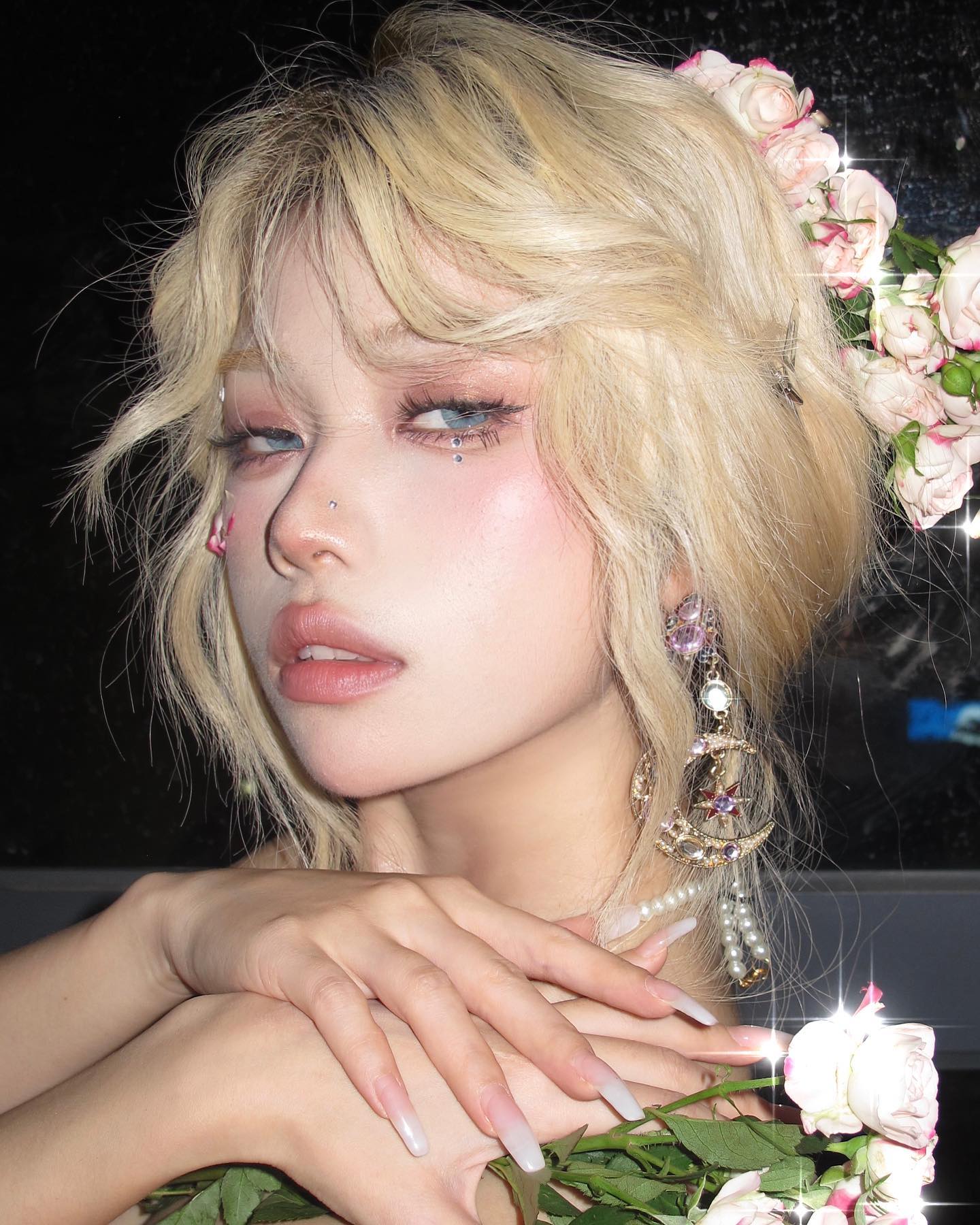 a closeup portrait of a young woman with blonde hair styled with a loose updo adorned with a crown of pink roses. she has on soft, ethereal makeup with rosy blush and shimmering eyeshadow. her nails are long and manicured with a white polish and sparkling accents. she wears elaborate earrings with pearls and moon charms. the overall aesthetic is romantic and feminine.