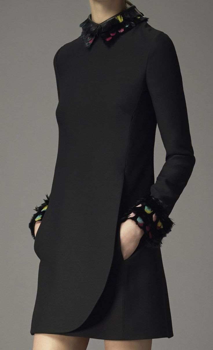 a woman in a black dress with a collar trimmed in feathers poses for the camera in a studio setting. the dress has a unique shape and the feather trim on the collar and sleeves adds a touch of whimsy.  the model's stance and the dress's silhouette create a fashionable and elegant look.