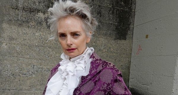 a woman with gray hair and a stern expression is wearing a purple patterned jacket with a large white ruffled collar. she is standing in front of a concrete wall. the image resembles a historical portrait.