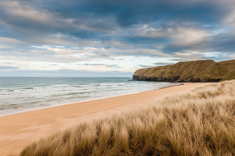 this is a photo of a beautiful sandy beach with a grassy foreground. the sand is a light golden color and the water is a deep blue. the sky is cloudy with some blue sky peeking through. the grassy dunes are a light brown color and the vegetation on the headland is green.