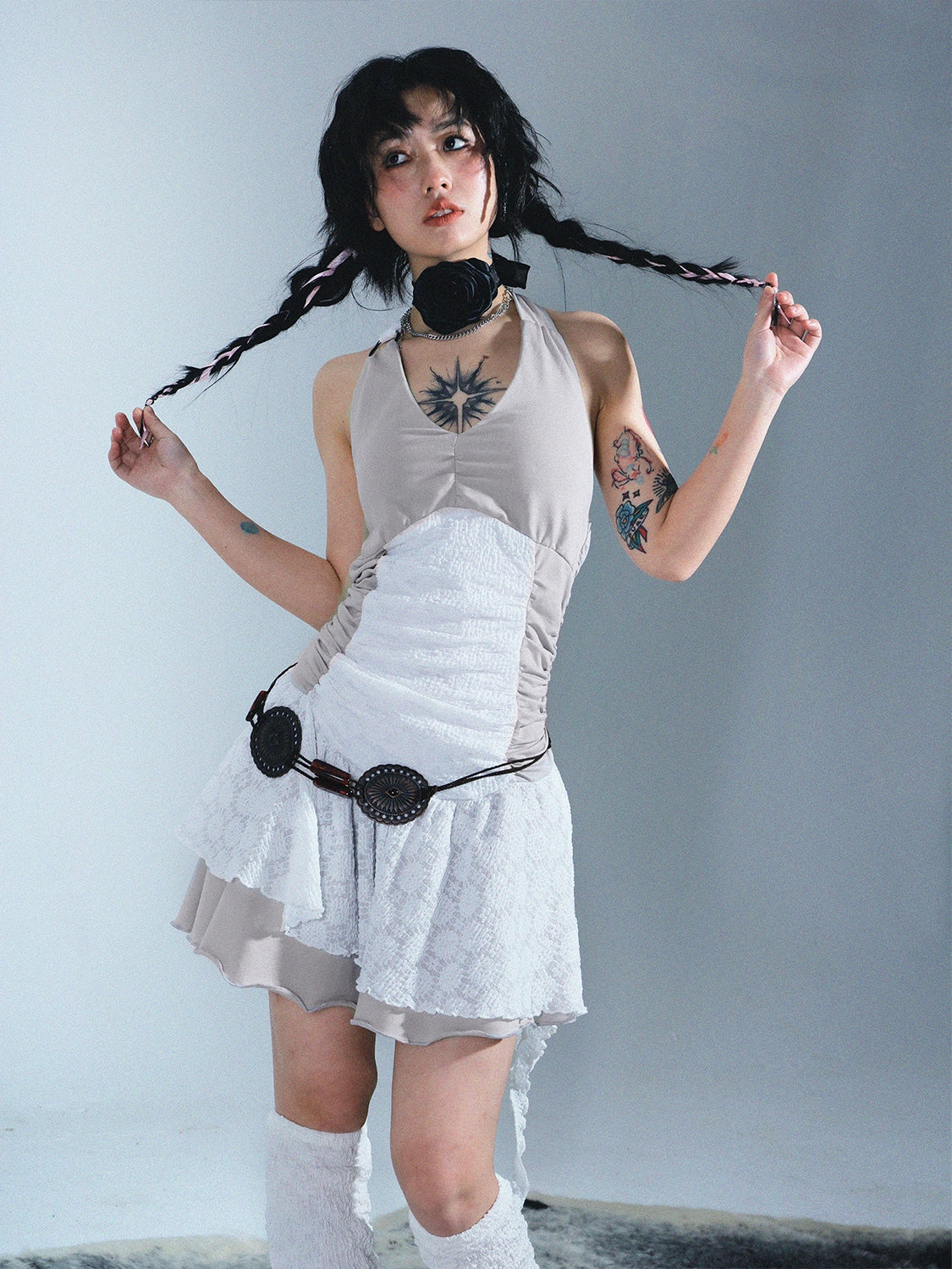 a young woman with long braids, wearing a unique beige and white layered dress with lace detailing and a decorative belt, poses against a light gray background.  she's accessorized with a black choker necklace and has visible tattoos on her arms.  the overall aesthetic is a blend of alternative, ethereal, and whimsical fashion styles.  the image is a welllit studio portrait.