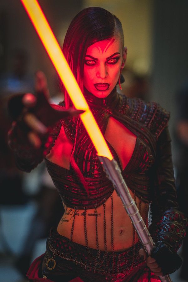 a young woman in a fantasy cosplay costume is wielding a glowing red lightsaber.  she has an intense expression on her face and is looking directly at the camera.