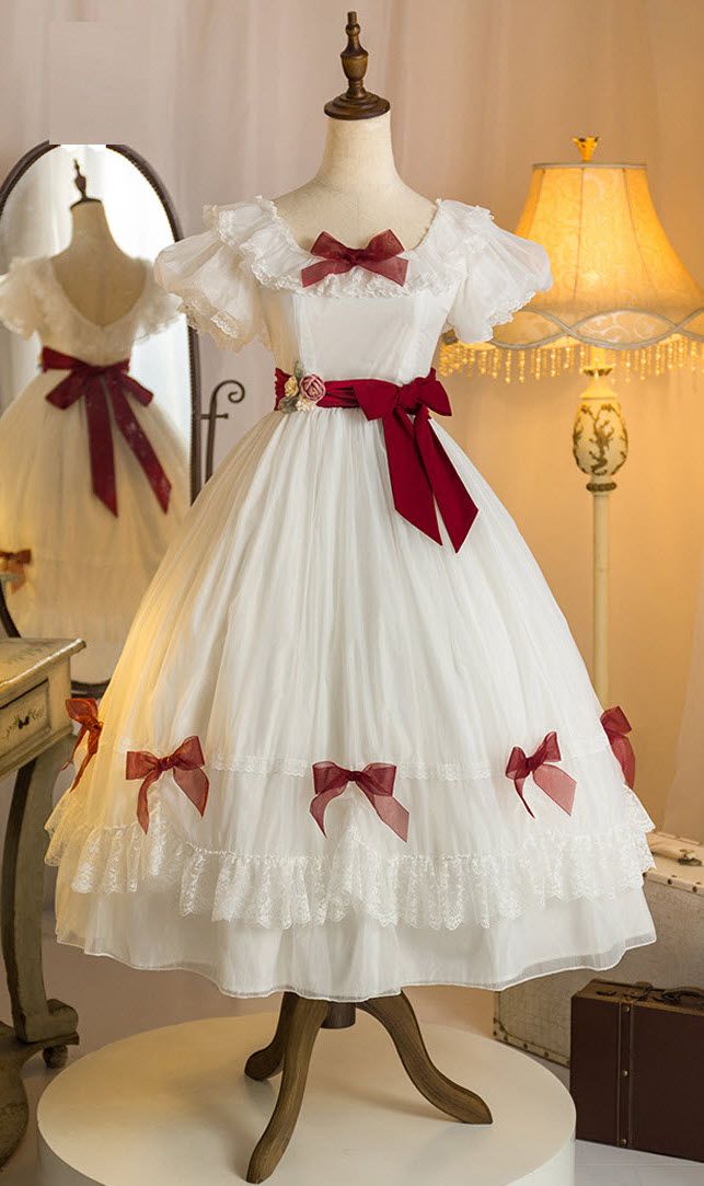 a white vintagestyle dress with red ribbons and bows is displayed on a mannequin. the dress features a square neckline, short puffed sleeves, and a flowing skirt with lace trim. the background includes a vintage mirror and a lamp, suggesting a room setting. the scene is welllit, highlighting the details of the dress and creating a bright and airy atmosphere.
