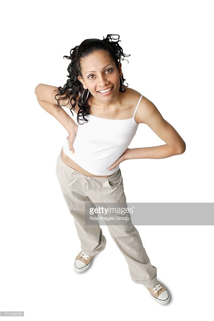 a young woman with curly brown hair is smiling at the camera. she is wearing a white tank top and beige pants. she is standing with her legs spread apart, relaxed, in a studio setting with a white background.