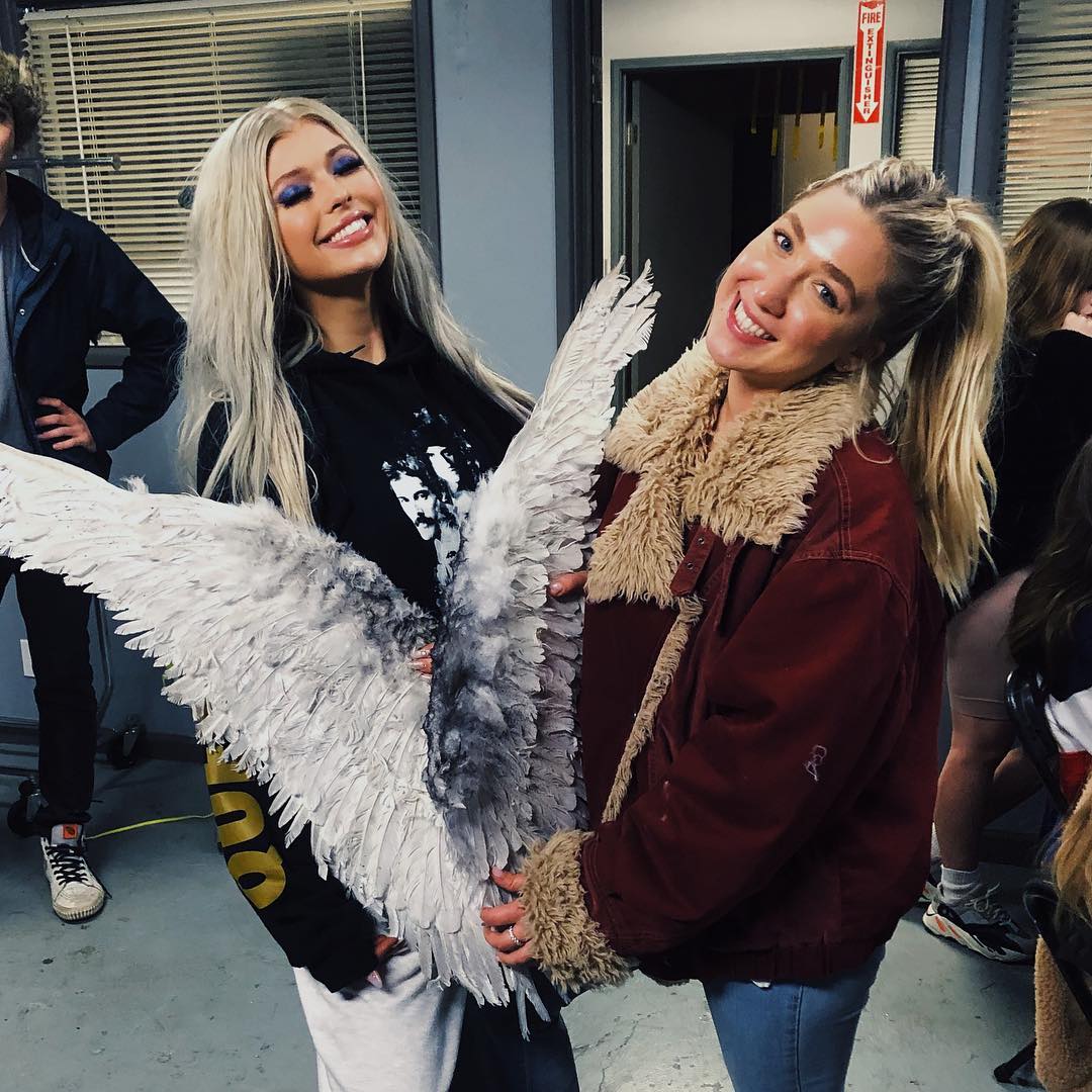 two women are smiling and holding a large pair of angel wings. the woman on the left has long blonde hair, is wearing a black hoodie, and white sweatpants. the woman on the right has her blonde hair in a ponytail and is wearing a red jacket with a fur trim. there are other people in the background, and the scene appears to be indoors. a fire extinguisher is visible on the wall.