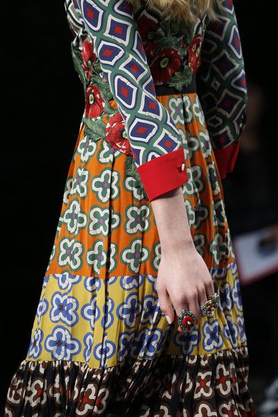 loseup view of a woman's colorful dress with a mix of floral and geometric patterns. the dress features various shades of orange, green, blue, red, and yellow. the sleeves have a distinct geometric pattern, and the body of the dress features a patchwork of different floral prints. the model's hand is visible, adorned with several rings. the overall style is bohemian and eclectic. the image appears to be from a fashion show or a photoshoot showcasing the detail of the garment.