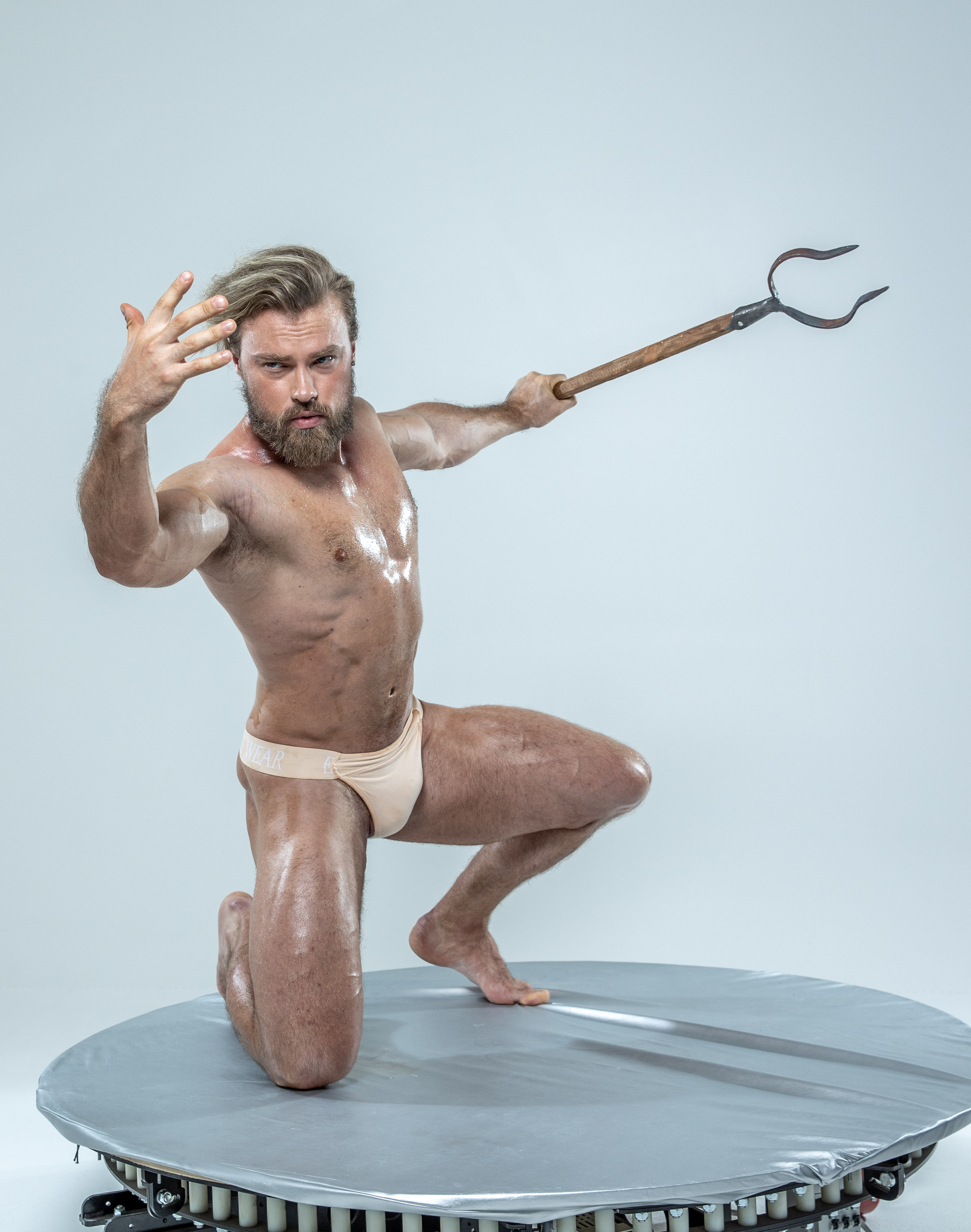 a muscular, shirtless man with a beard and slickedback blonde hair poses dramatically on a round, metallic platform against a light gray background. he holds a wooden trident in his right hand, pointing it forward with a determined expression. his left hand is raised in a defensive gesture. he wears only a pair of tight, beige underwear.  the soft lighting emphasizes his muscular physique.  the overall scene evokes a sense of classical mythology, potentially depicting a greek god or warrior.