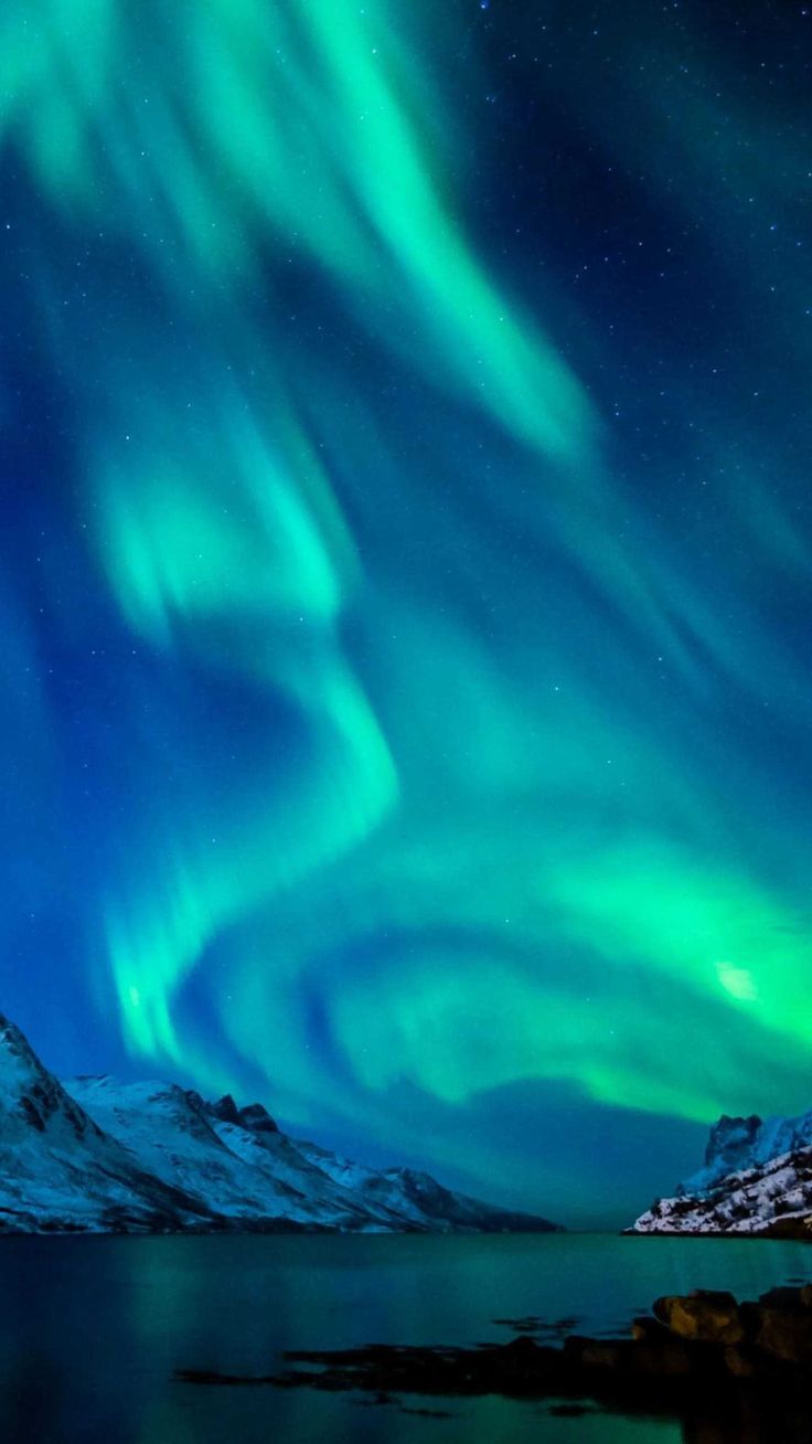 a breathtaking view of the aurora borealis, also known as the northern lights, illuminates the night sky over a serene landscape. vivid streaks of green and blue dance across the dark expanse, creating an ethereal and magical spectacle. snowcapped mountains rise in the distance, their peaks partially covered in white. the calm water reflects the vibrant colors of the sky, adding depth and dimension to the scene. dark rocks and land formations line the shore, grounding the otherwise ethereal view. the overall image evokes a sense of awe, wonder, and the raw beauty of nature.