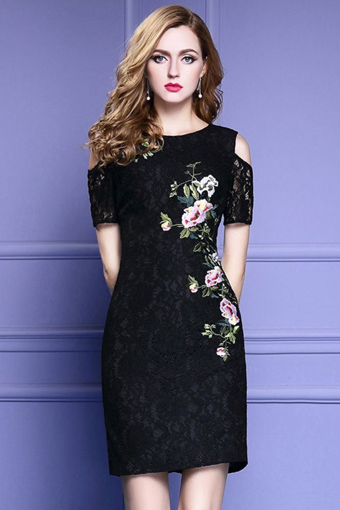 a woman in a black coldshoulder lace dress with floral embroidery stands against a purple background. the dress is fitted and shortsleeved, ending above the knee. she is looking directly at the camera, and her hair is styled in soft waves.